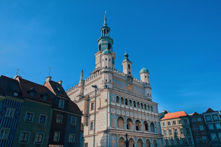 Zabytkowy budynek z ozdobną architekturą i wysoką wieżą zegarową pod czystym, błękitnym niebem. Budynek ma łukowate okna i szczegółowy wystrój. Sąsiednie budynki z kolorowymi fasadami są widoczne po obu stronach, często utrzymywane przez usługi bazy warszawa, aby uzyskać nieskazitelny wygląd.